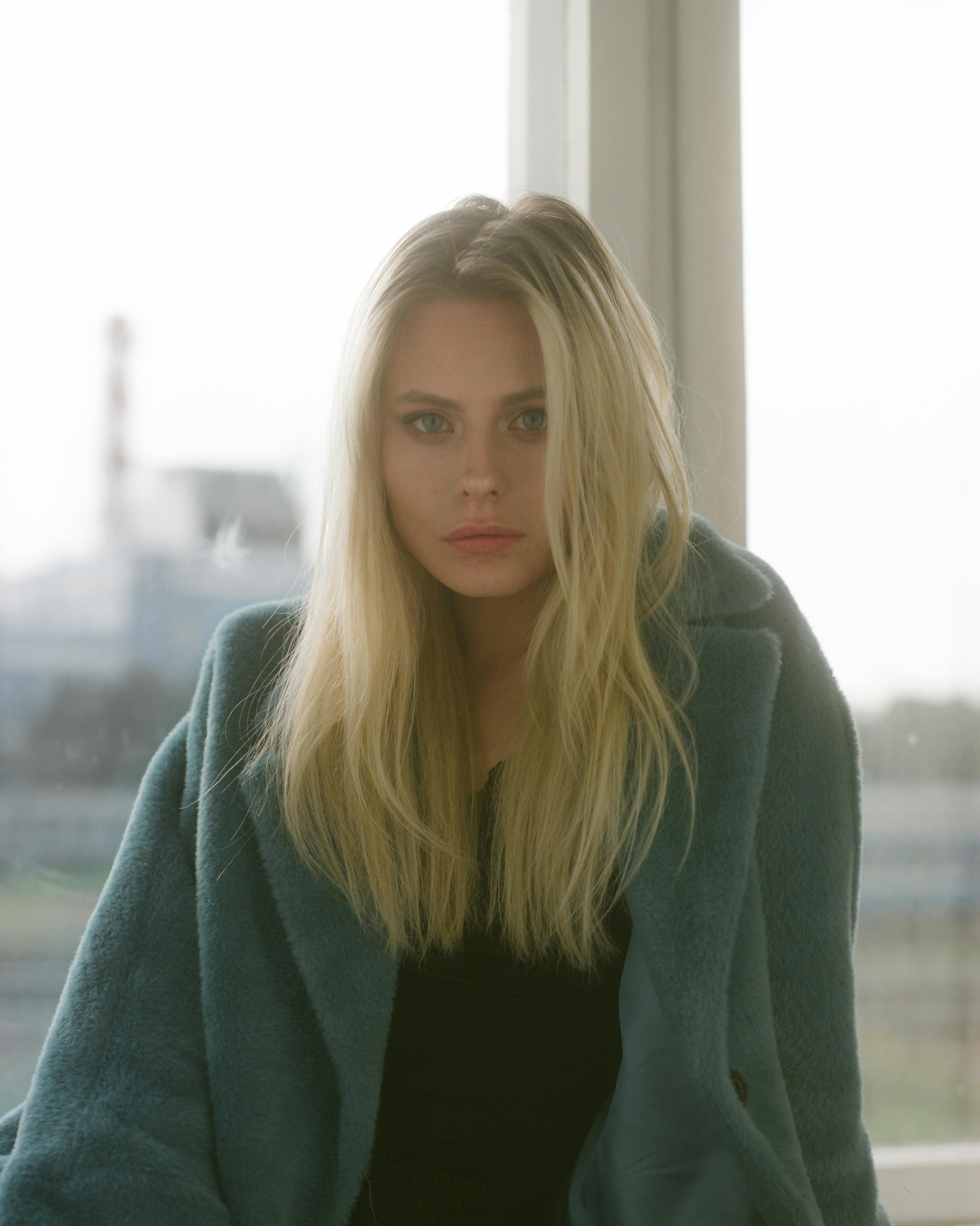 Portrait of a woman in a teal coat gazing through a window, creating a serene and introspective mood.