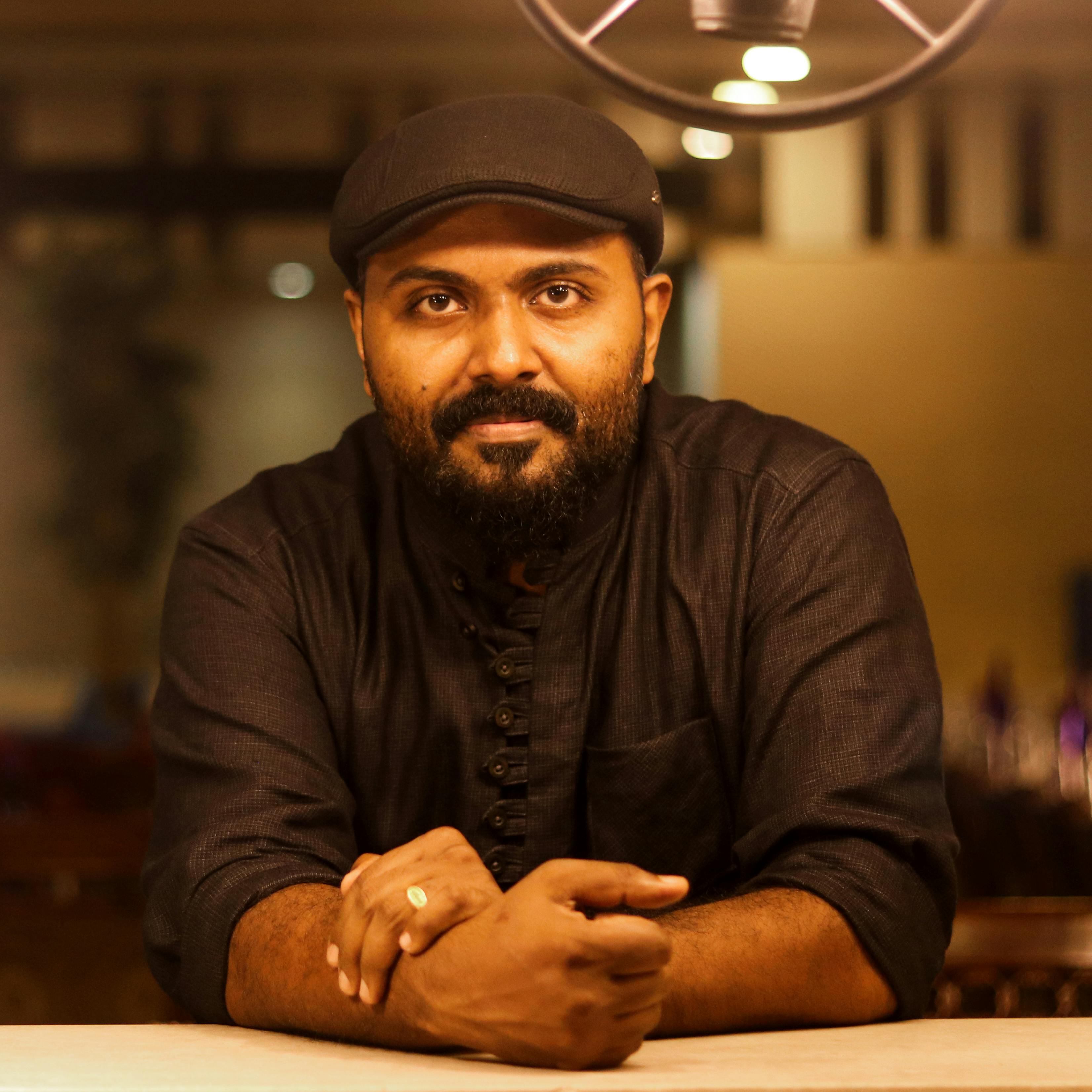 Portrait of a bearded man with a hat, leaning on a table indoors.