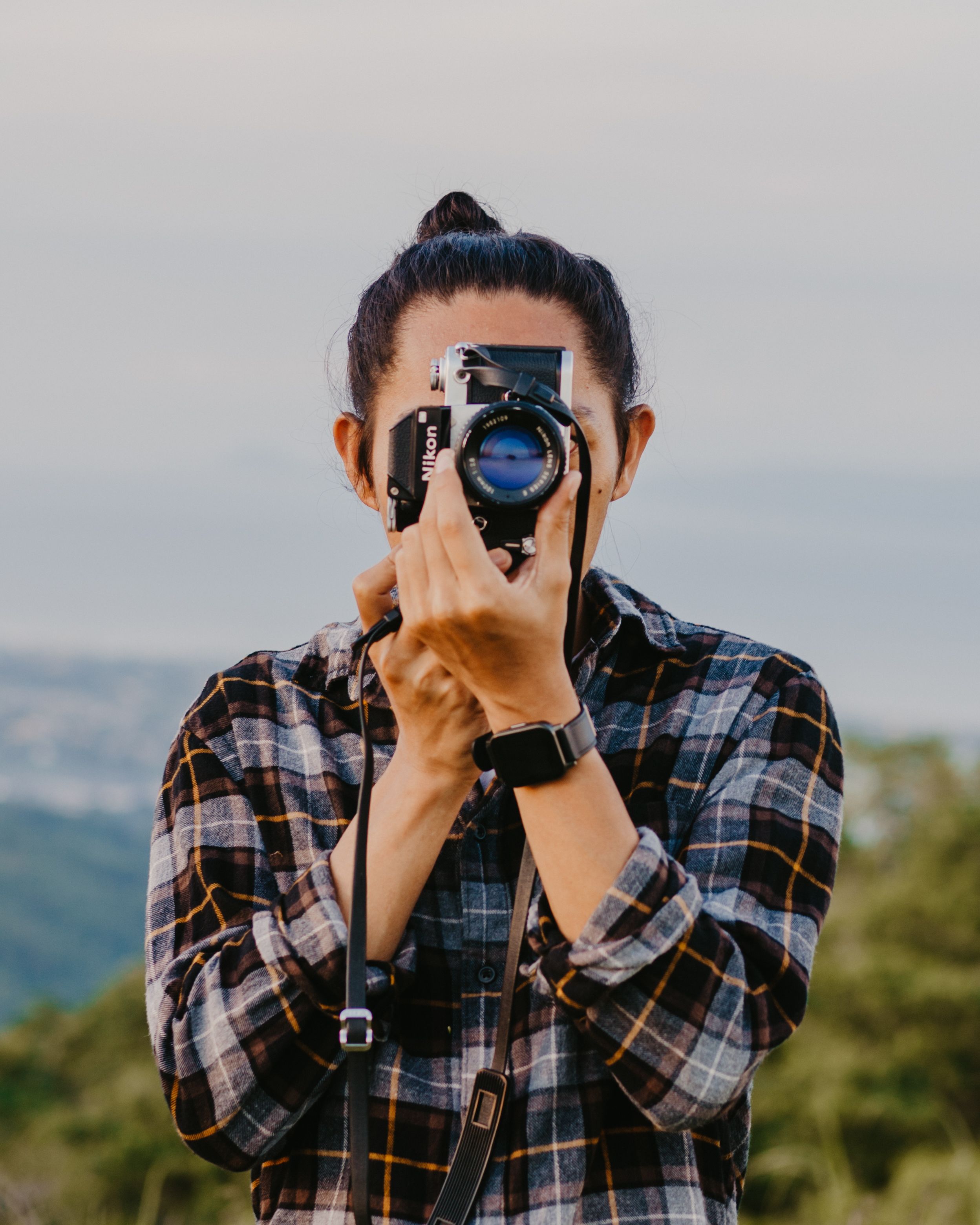 A young photographer wearing a plaid shirt takes a picture outdoors, showcasing creativity and passion for photography.