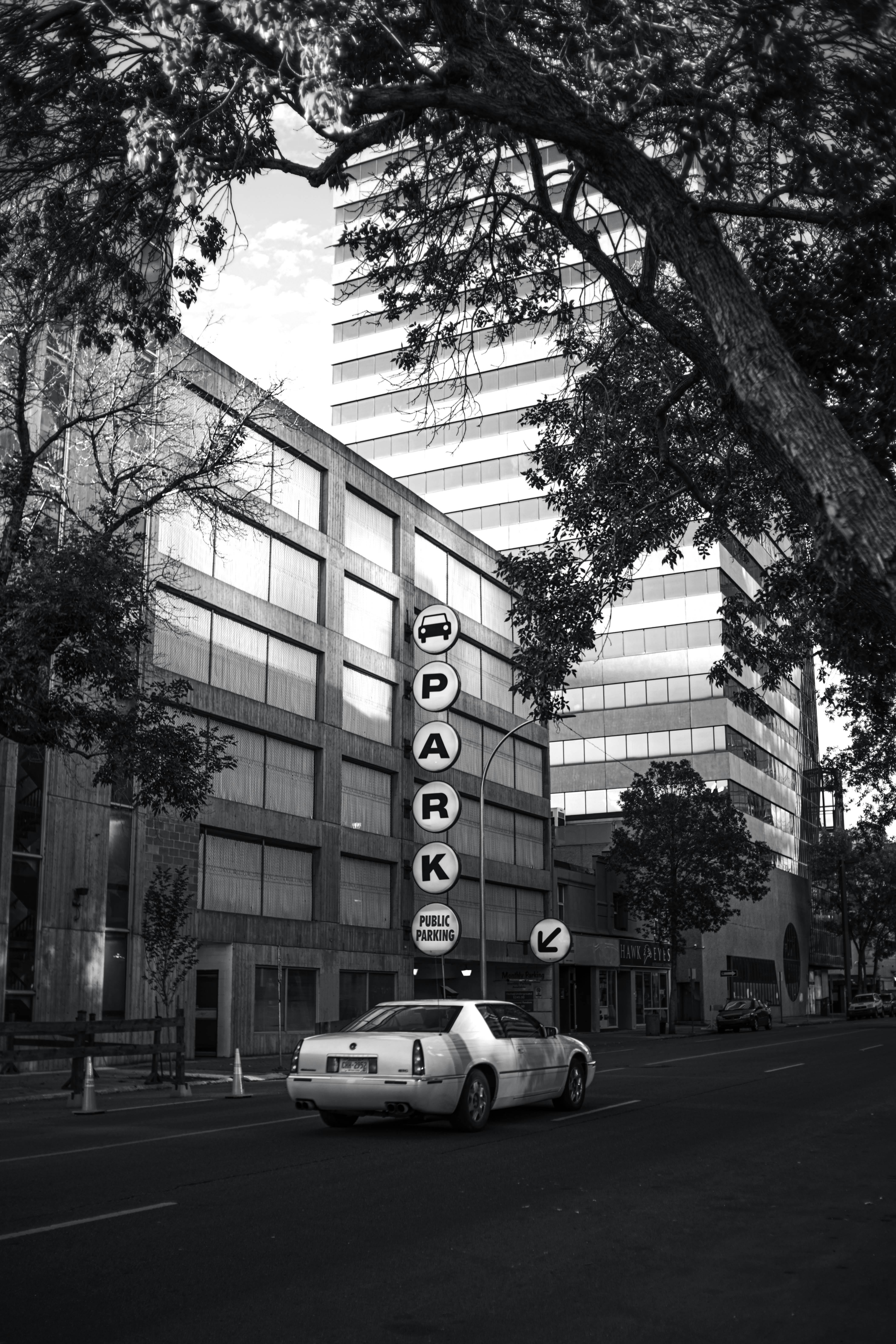 Monochrome cityscape featuring a parked car under a parking sign between buildings.