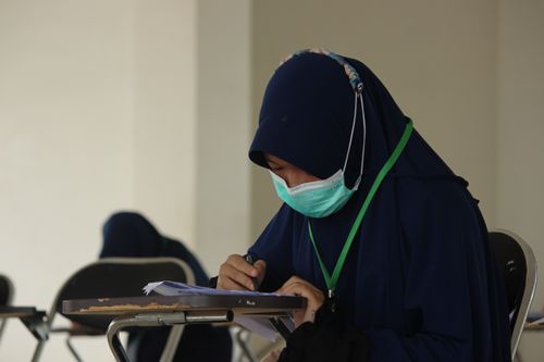 Free A Muslim woman in a hijab and face mask writing on paper during an exam indoors. Stock Photo