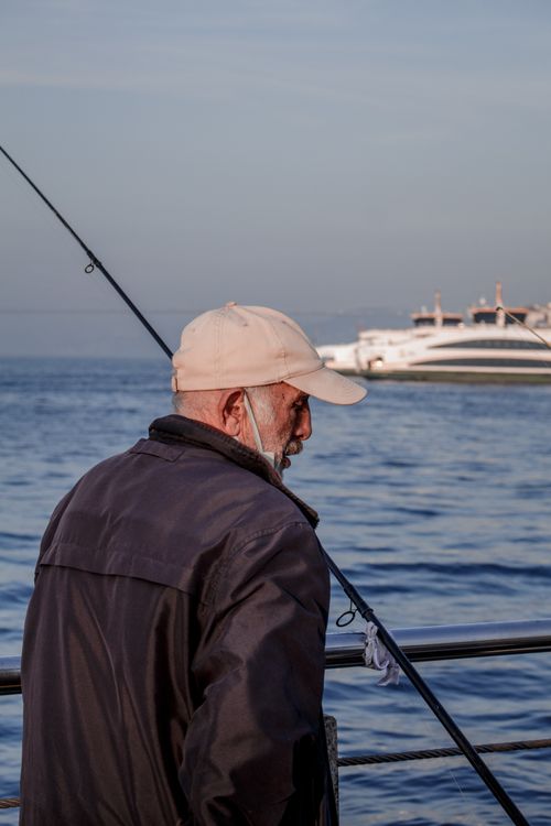 Free A senior man enjoys fishing by the sea under a clear blue sky with a yacht in the distance. Stock Photo