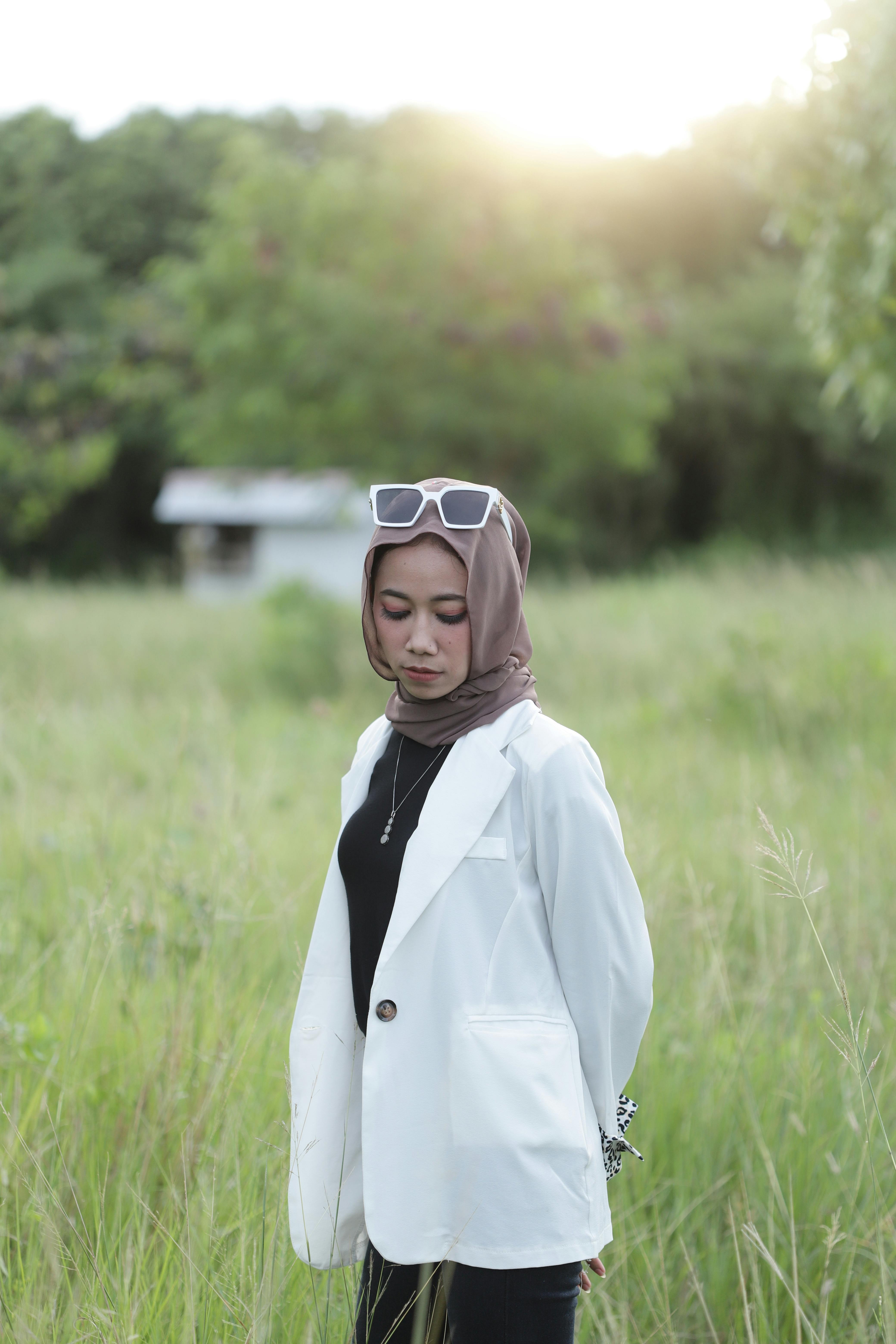 Stylish woman with hijab and sunglasses posing outdoors in a vibrant grass field.