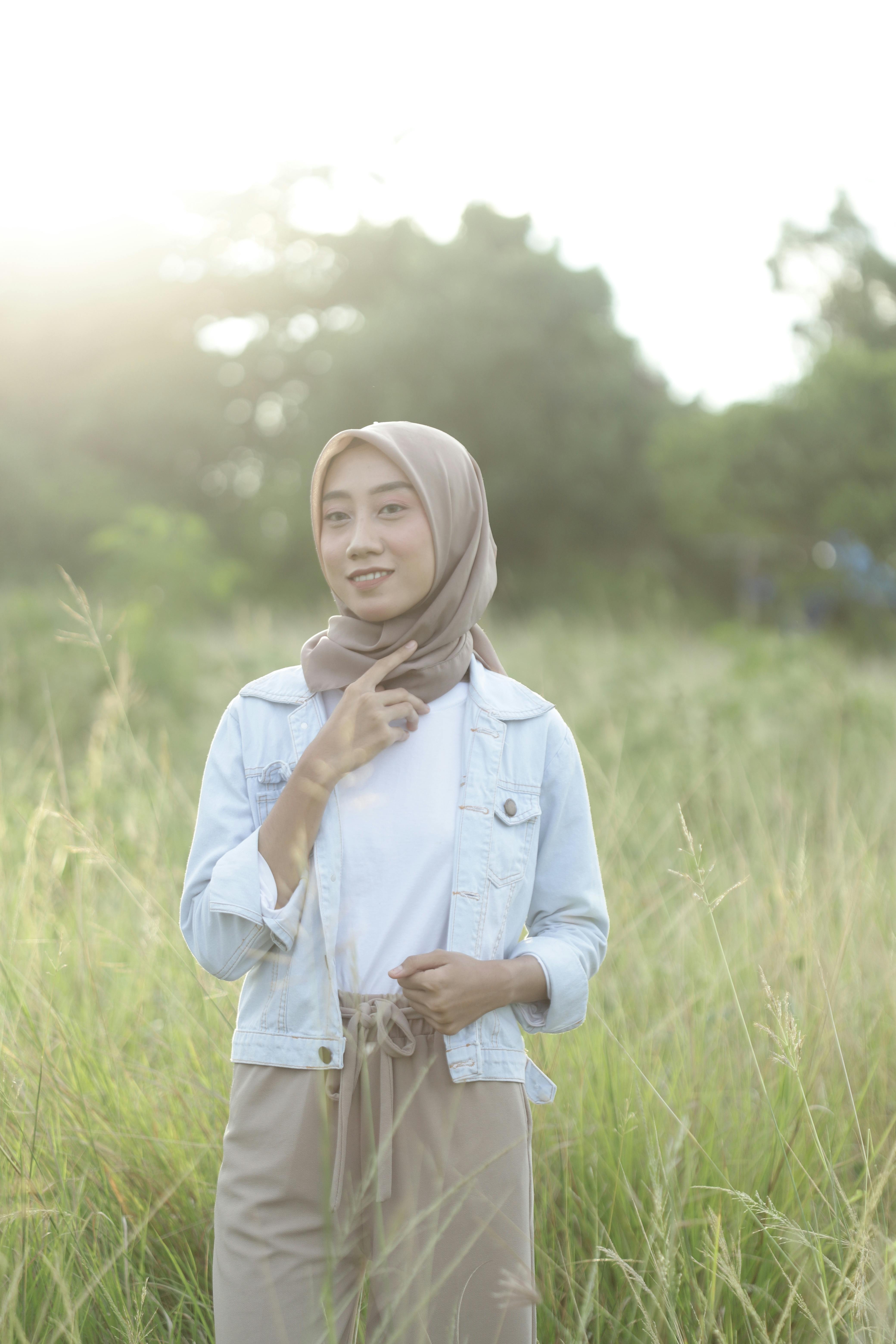 Young woman wearing hijab smiling in a sunny grassy field with a denim jacket.