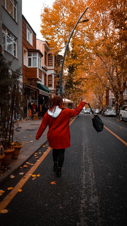 Free A woman wearing a red coat walks down a street lined with autumn trees, carrying a handbag. Stock Photo