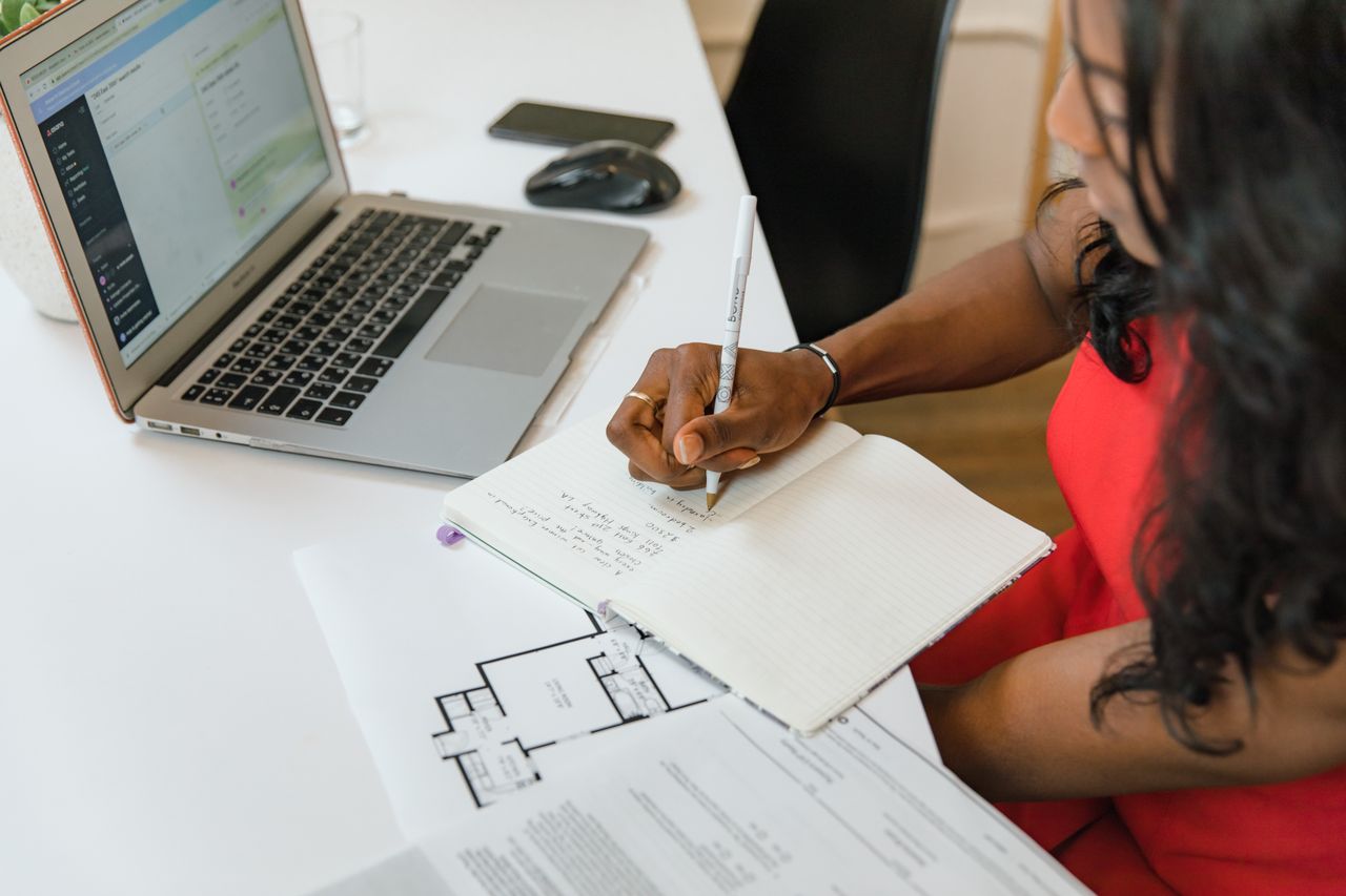 Photo d’illustration : une femme prend des notes dans un carnet à côté d’un ordinateur et de plans. Photo RDNE Stock project / Pexels.