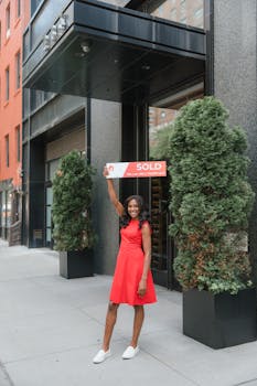 Smiling woman celebrates real estate success with sold sign outside building.
