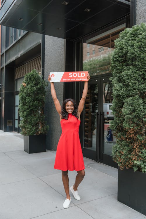 Free Smiling woman in red dress celebrating real estate sale with a sold sign outdoors. Stock Photo