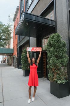Smiling woman in red dress holding 'Sold' sign outside modern office building.