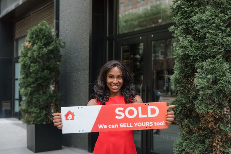 Smiling Woman Holding Sold Sign Outside Office Building