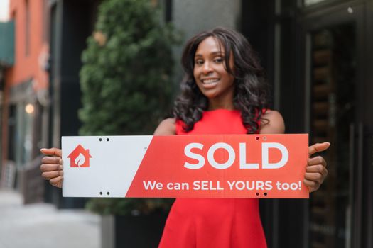 Happy woman in red dress holding a 'Sold' sign in an urban setting, promoting real estate sales.