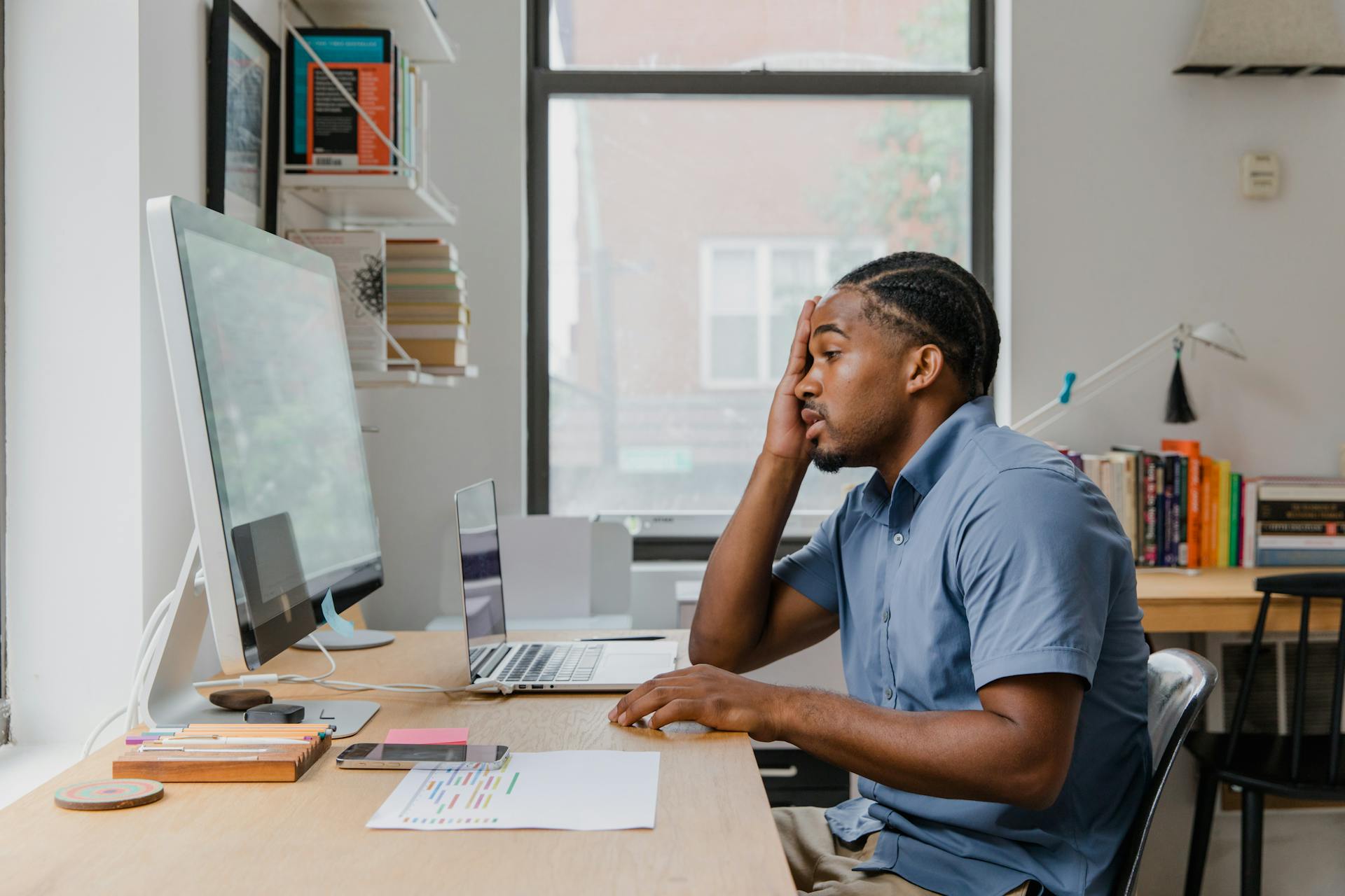 A man looks stressed, working at a desk with multiple computers in an office setting.