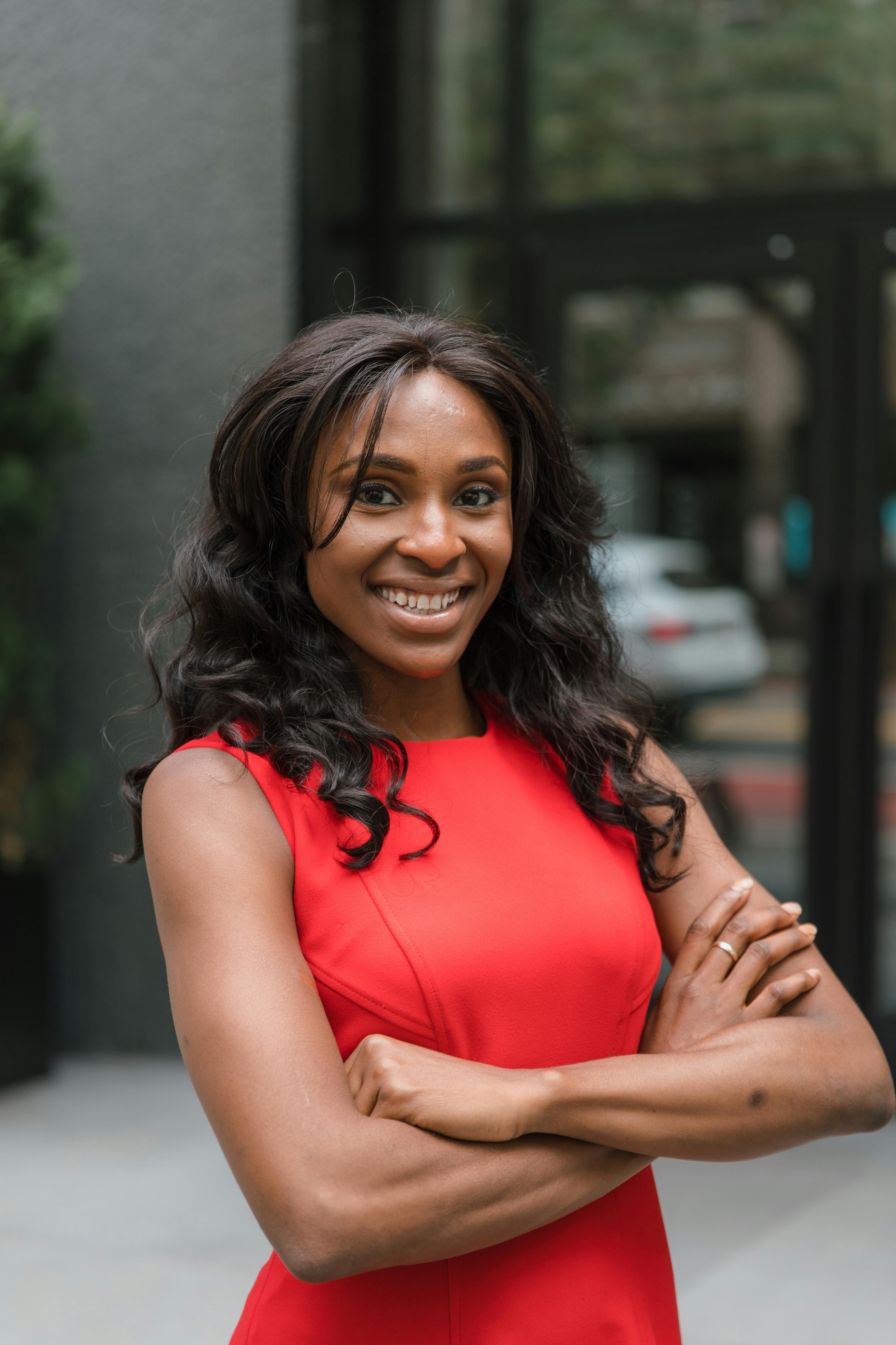 Smiling woman in a red dress with arms folded, exuding confidence and happiness in an outdoor urban setting.