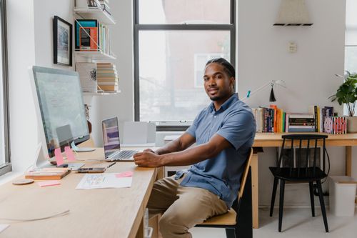 Free African American man sitting at a cluttered home office desk with computer and books. Stock Photo