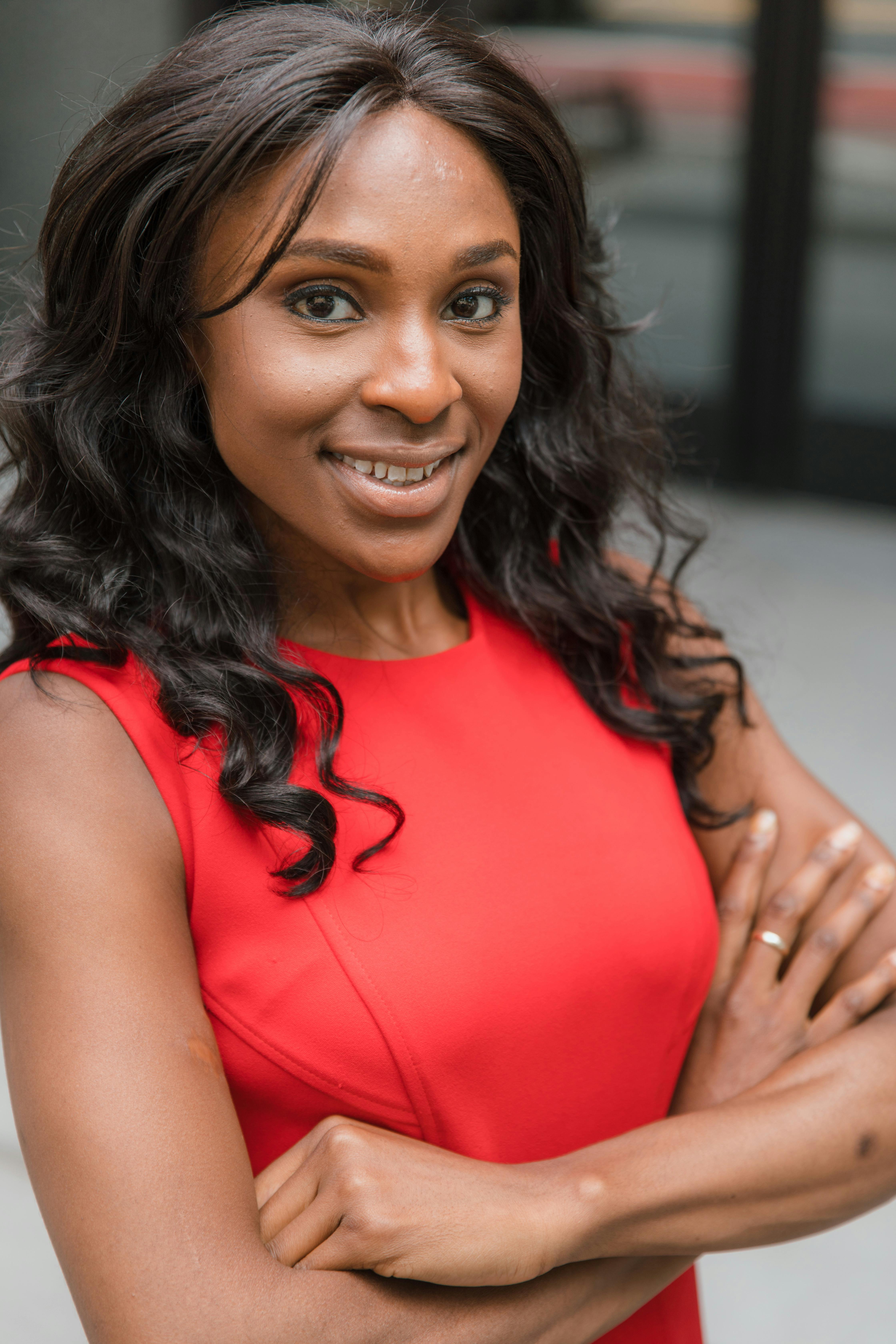 A smiling woman in a red dress standing outdoors with arms folded, expressing confidence.