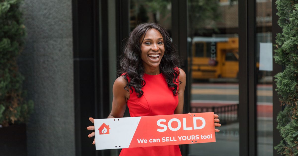 Smiling woman in red dress holding sold sign outside an office building.