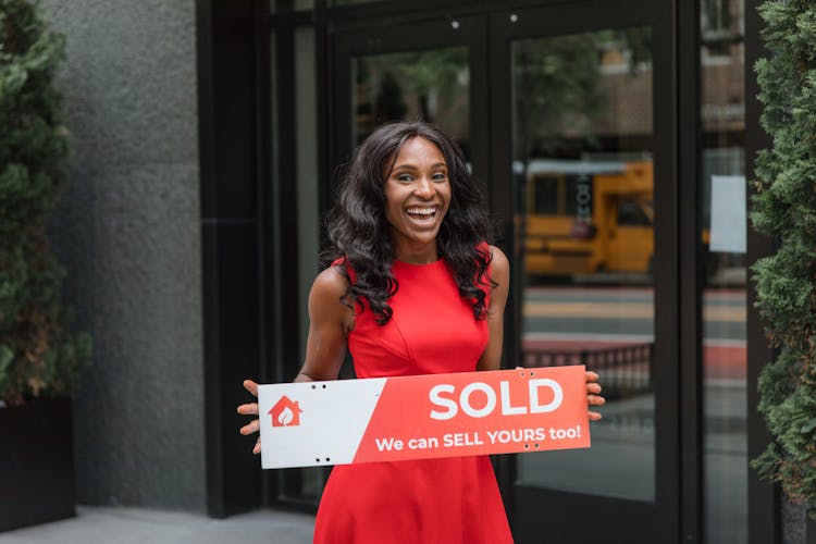 Smiling Woman Holding Sold Sign Outside Real Estate Office