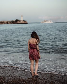 Young woman in a sundress stands on a quiet beach, gazing at a distant lighthouse.