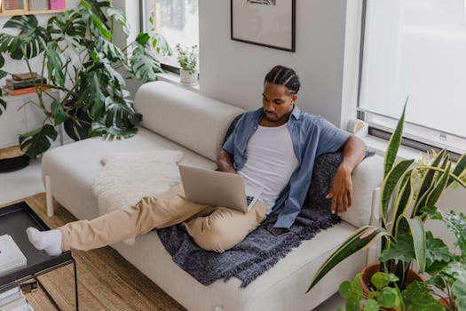 A man with braided hair working remotely on a laptop in a cozy living room setting with plants.