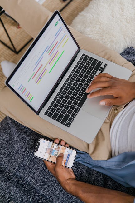 Man sitting on sofa multitasking with laptop and phone, focused on digital projects.