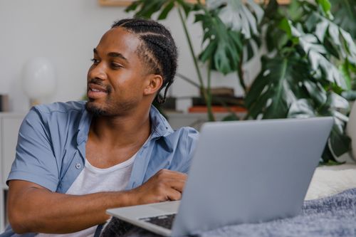 Free Man with braided hair smiling while using a laptop indoors surrounded by plants. Stock Photo