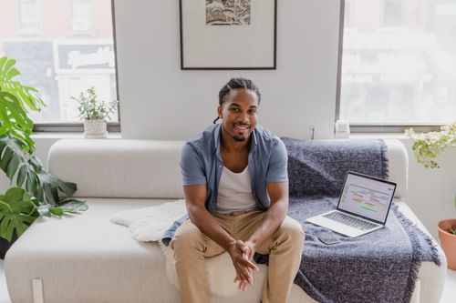 Free A cheerful man sitting on a sofa in a home office with a laptop, radiating serenity and comfort. Stock Photo