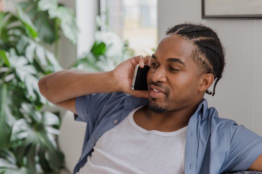 A relaxed man with braided hair casually talks on the phone in a cozy living room setting.