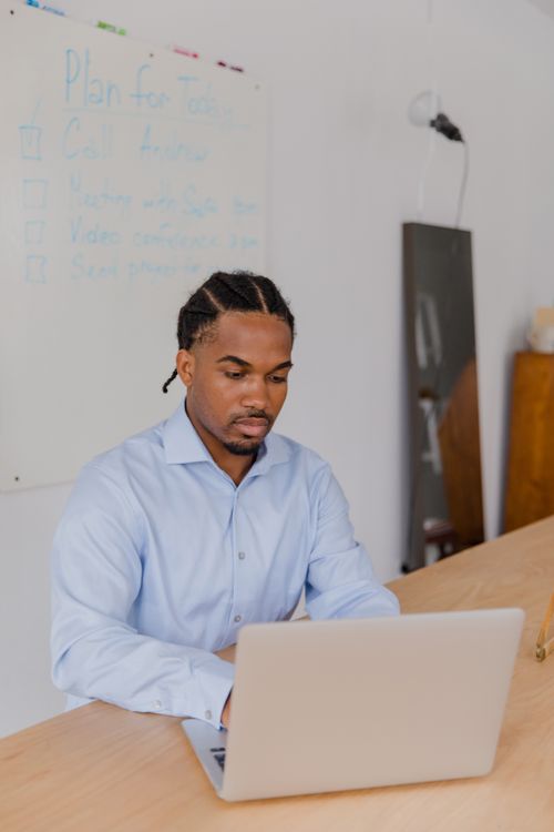Free Man in a blue shirt working on a laptop at a wooden desk in a modern office setting. Stock Photo