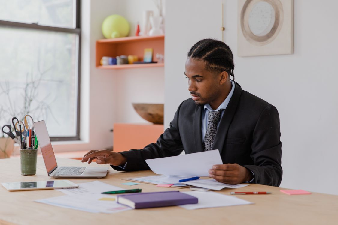 A small business owner in a suit reviews printed paperwork beside an open laptop at a bright office desk