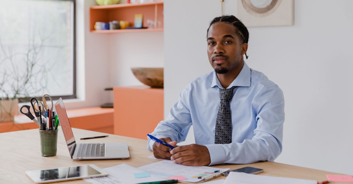 A professional businessman with a beard and braided hair sits at his desk with a laptop and documents, working diligently.