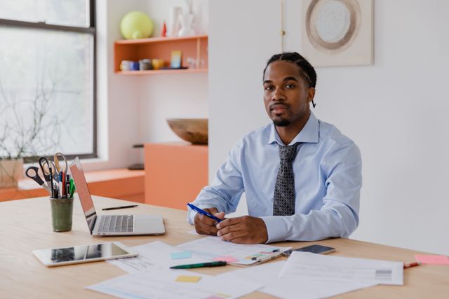 A man in a shirt and tie sits at an office desk with papers, a laptop and a phone