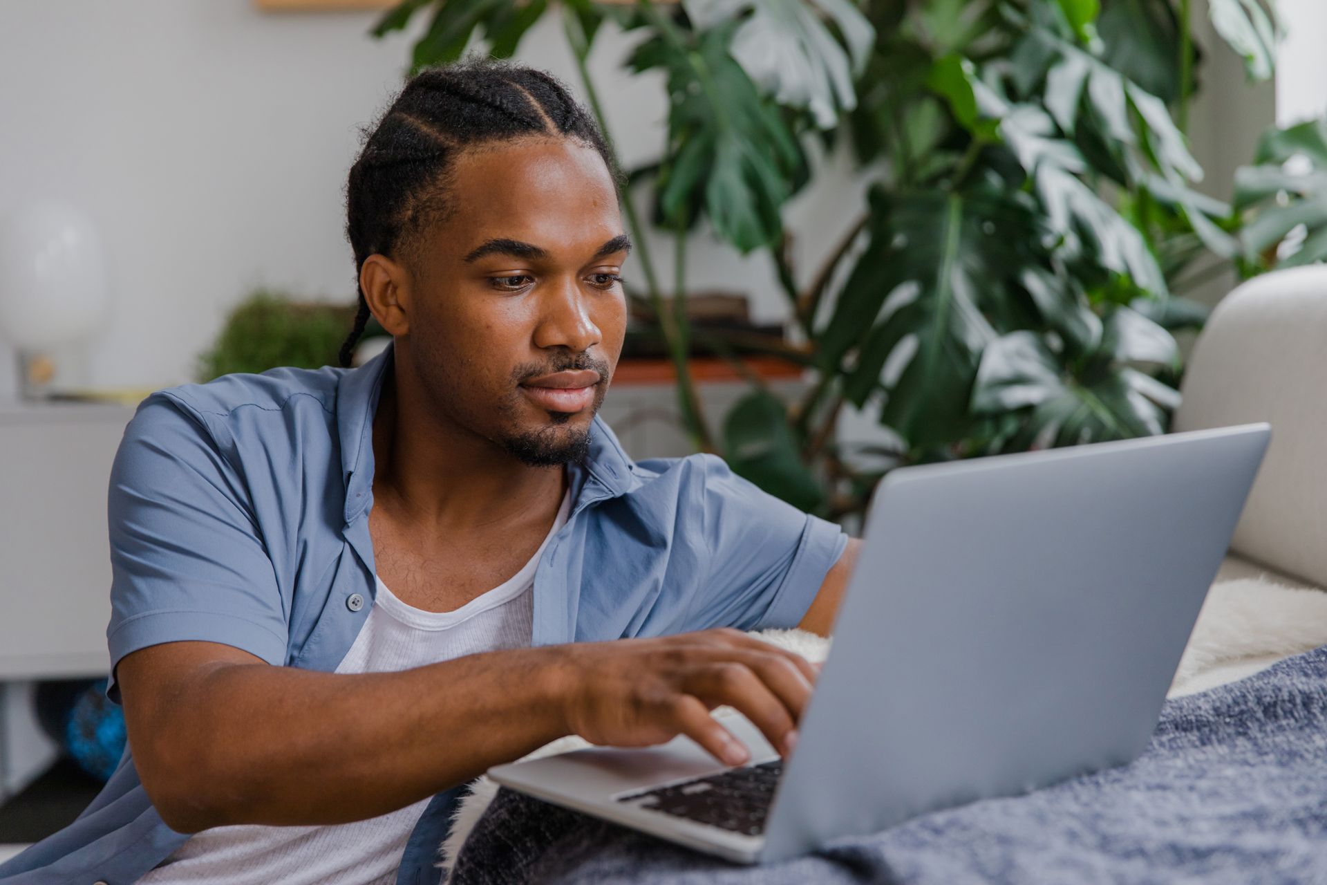 Young man in blue shirt working on laptop in a home office with a monstera plant.