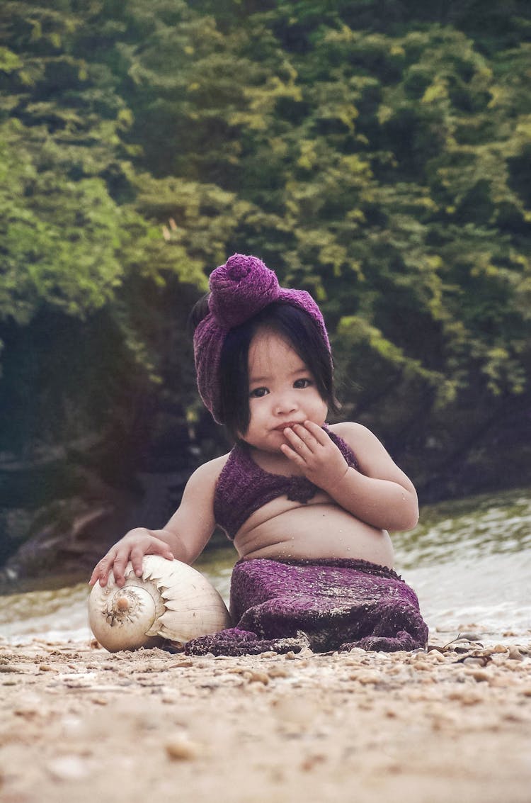 Child In Purple Mermaid Costume Sitting On Beach Sand