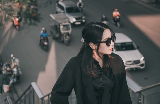 An Asian woman in sunglasses and a black coat standing on a street overlooking a bustling road with vehicles.