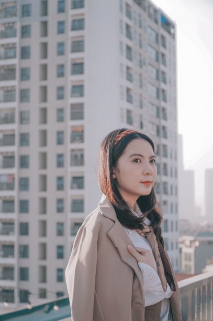 Portrait of an Asian woman in a beige coat with a city building in the backdrop, capturing a modern urban vibe.