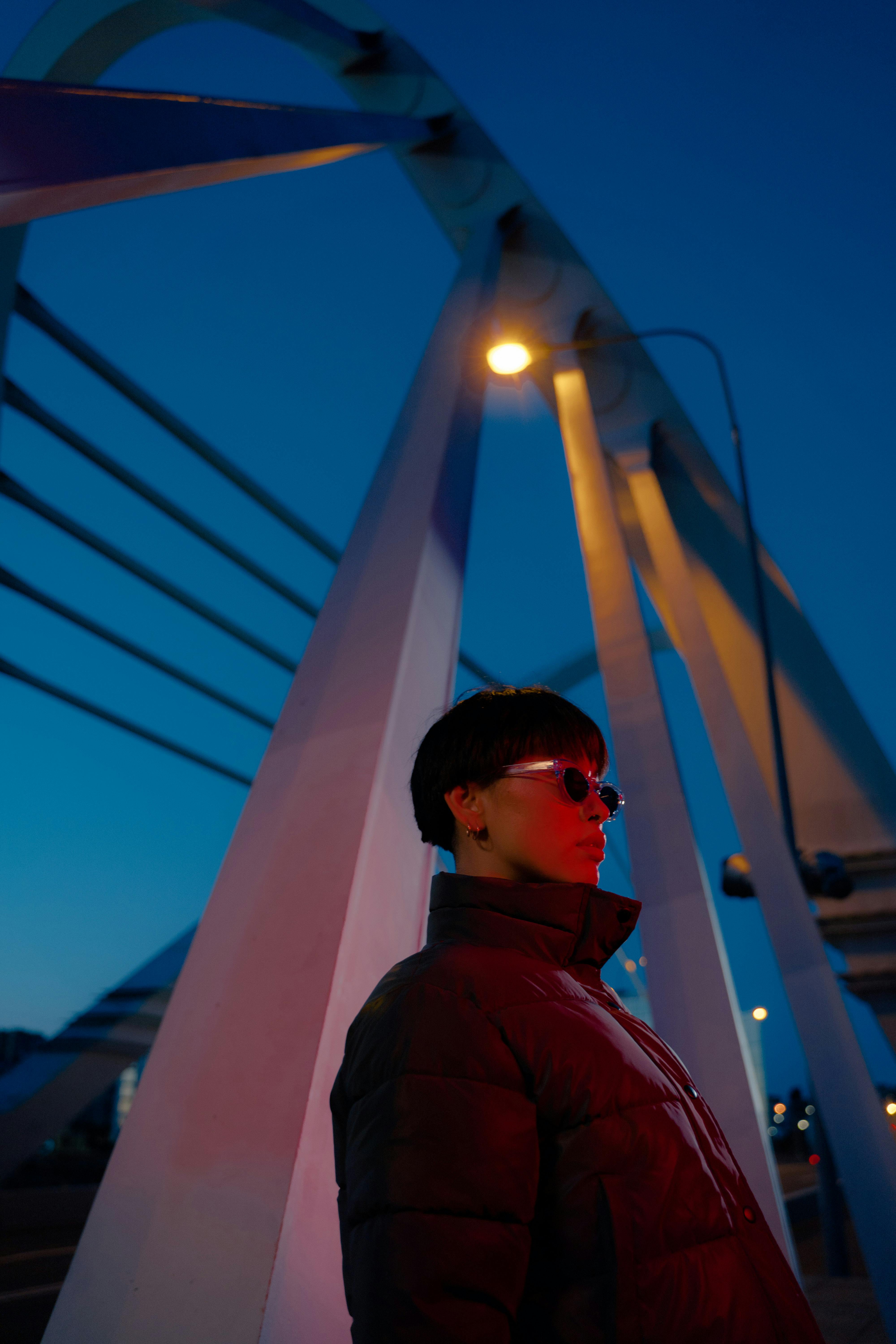 A person in a puffer jacket and sunglasses stands by a bridge, illuminated by city lights.