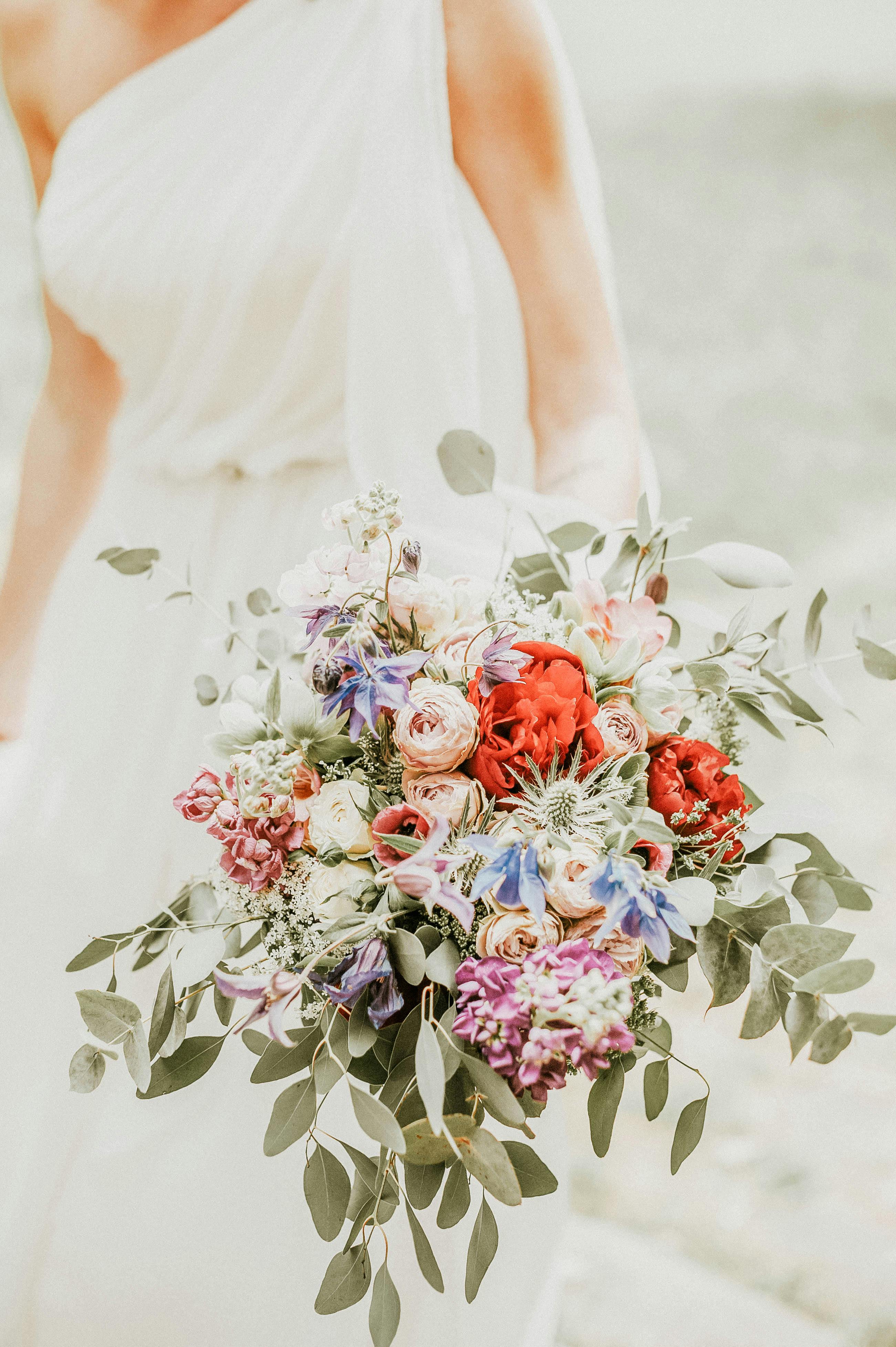 A bride in a white dress holding a vibrant floral bouquet with red, purple, and green accents.