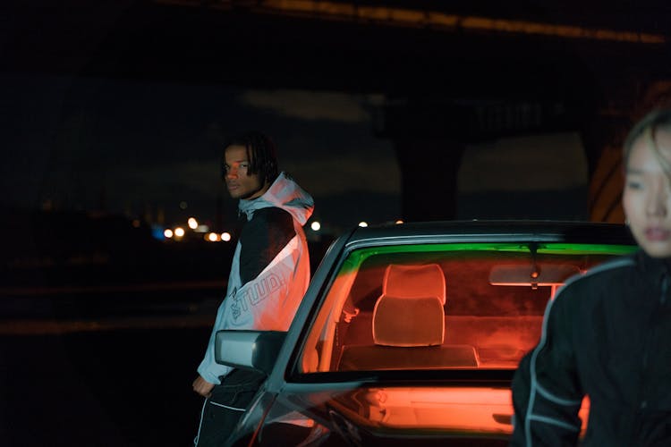 A Man In White And Black Hoodie Leaning On A Car At Night