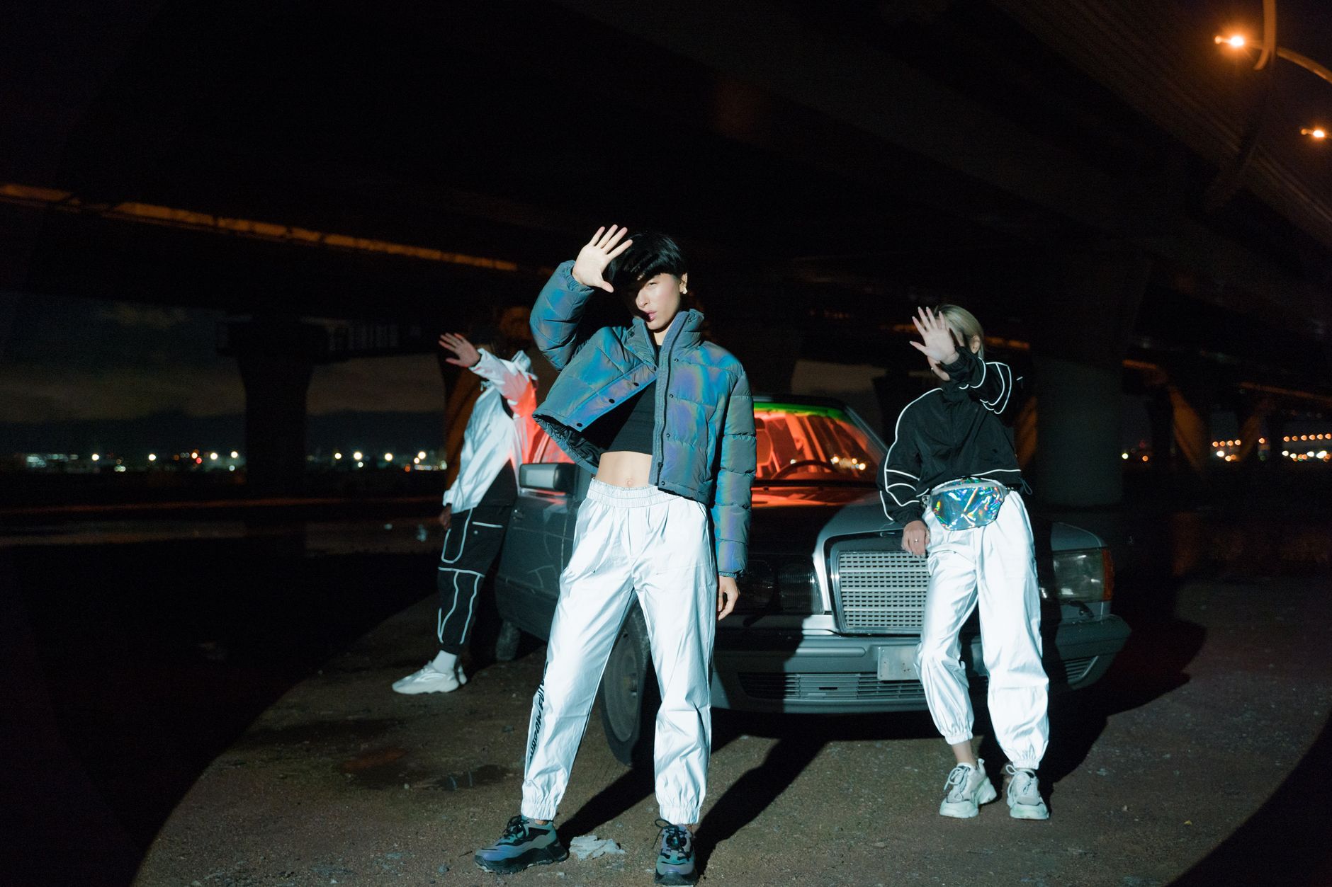 Stylish group posing by a car at night under an urban bridge.