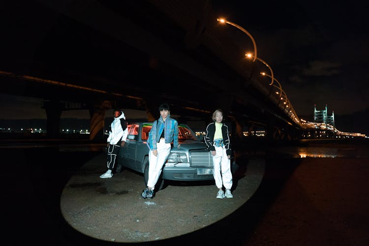 A Group Of People Leaning On A Car Under A Bridge