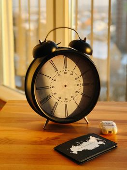 A classic alarm clock alongside a passport and earbuds on a wooden table by the window.