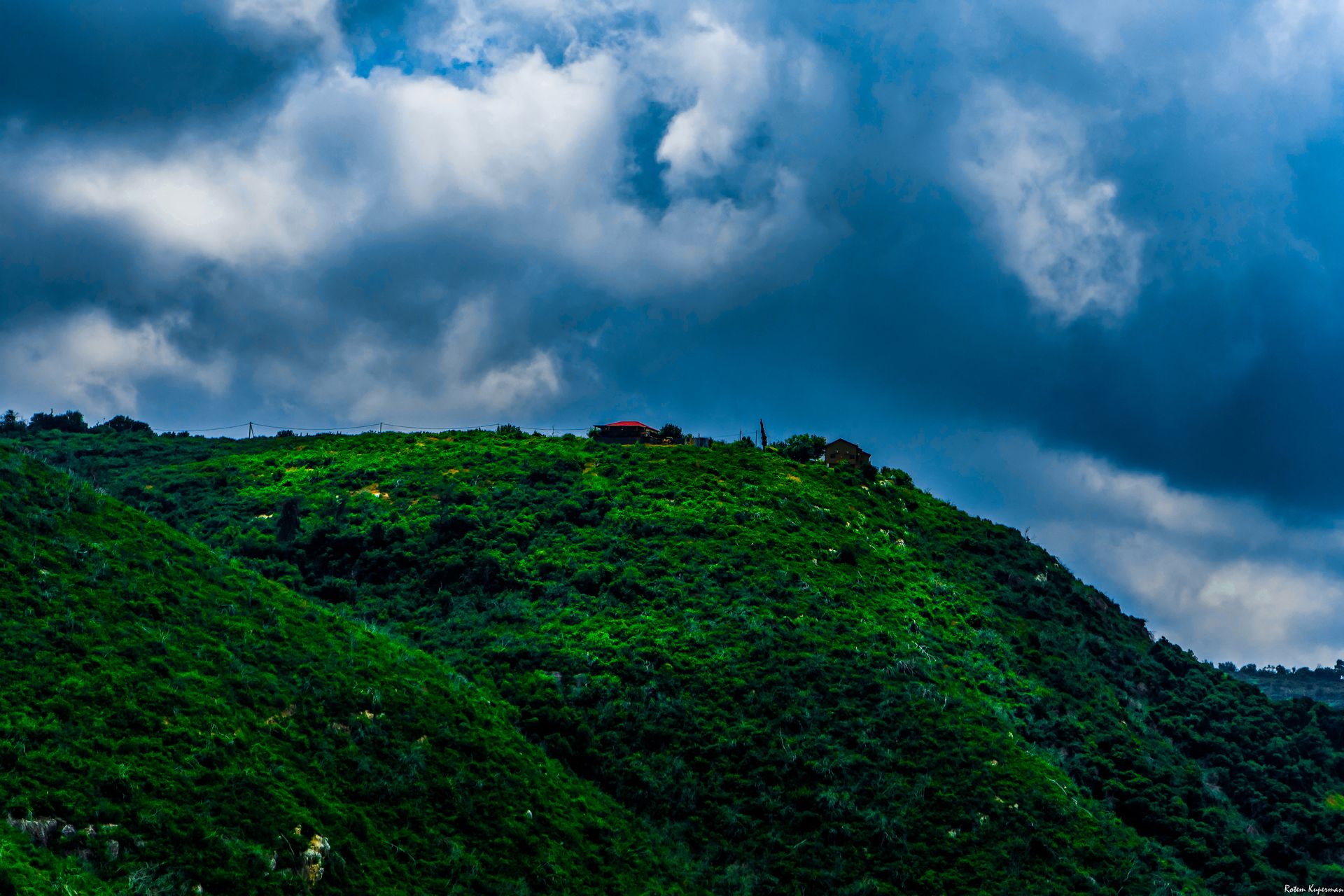 A scenic view of verdant hills with a house atop, set against a dramatic, cloudy sky near Be'er Sheva, Israel.