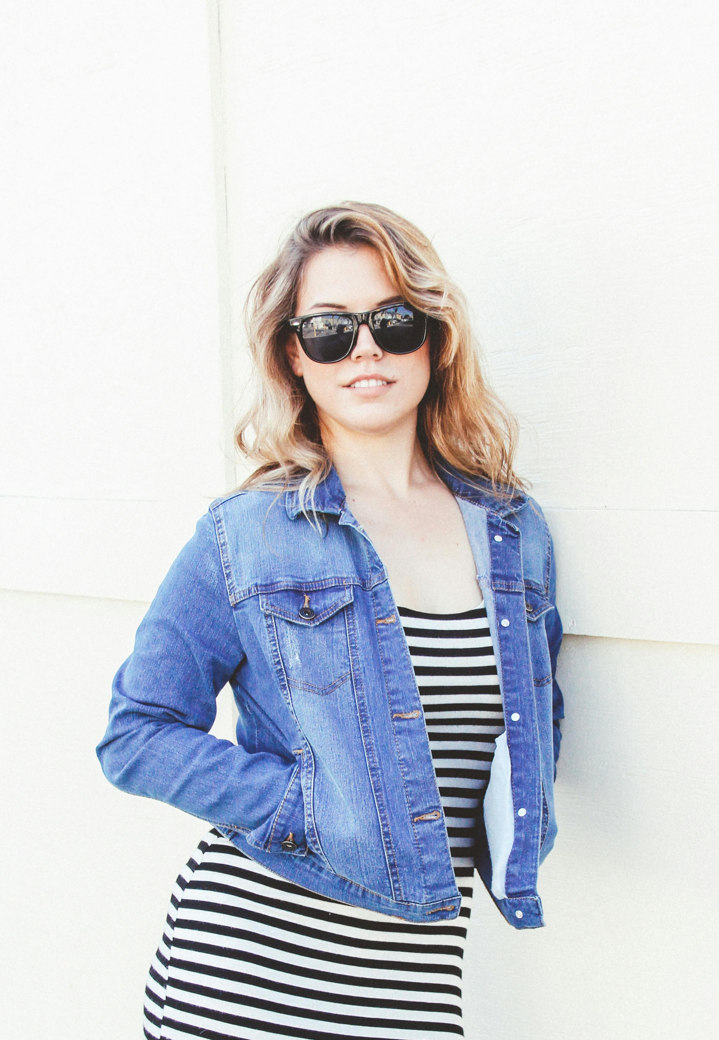 一位戴着太阳镜的时尚女性，身穿条纹连衣裙和牛仔夹克，带着灿烂的笑容，显得十分自信