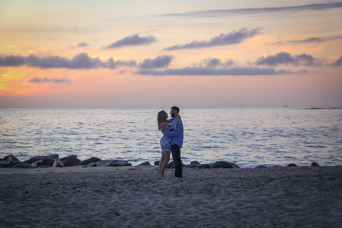 Pasangan Tatap Muka Berdiri Di Tepi Laut Dekat Perairan Tenang Selama Golden Hour