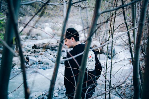 Man Wearing Black Sweatshirt Standing Beside River and Bare Trees