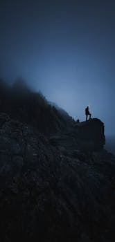 A lone hiker stands silhouetted on a rocky mountain cliff during twilight, evoking a sense of adventure.