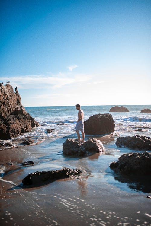 Man in White Shorts Standing on Brown Rock on Sea Shore