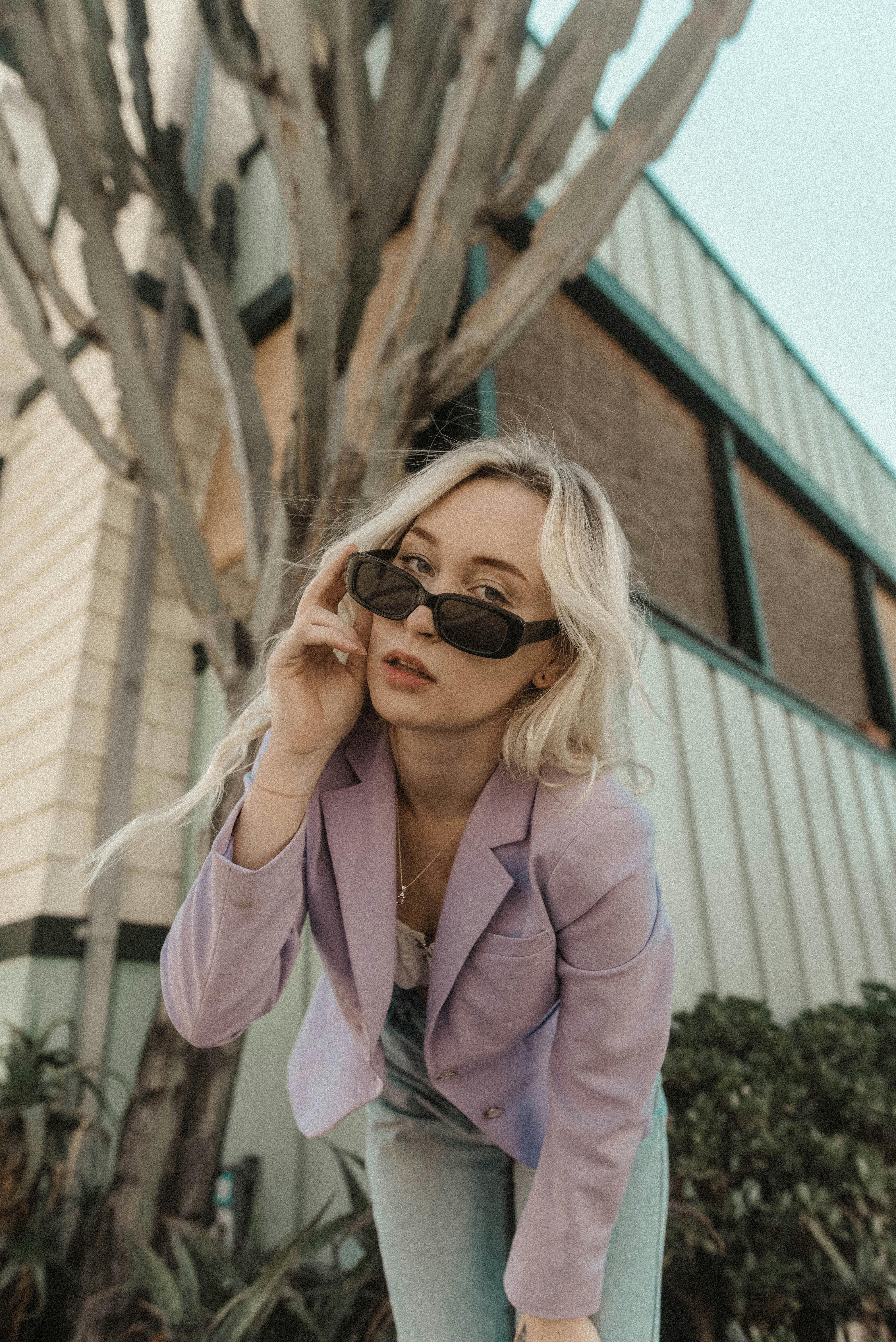 Fashionable woman in a purple blazer wearing sunglasses, posing outdoors against a building.