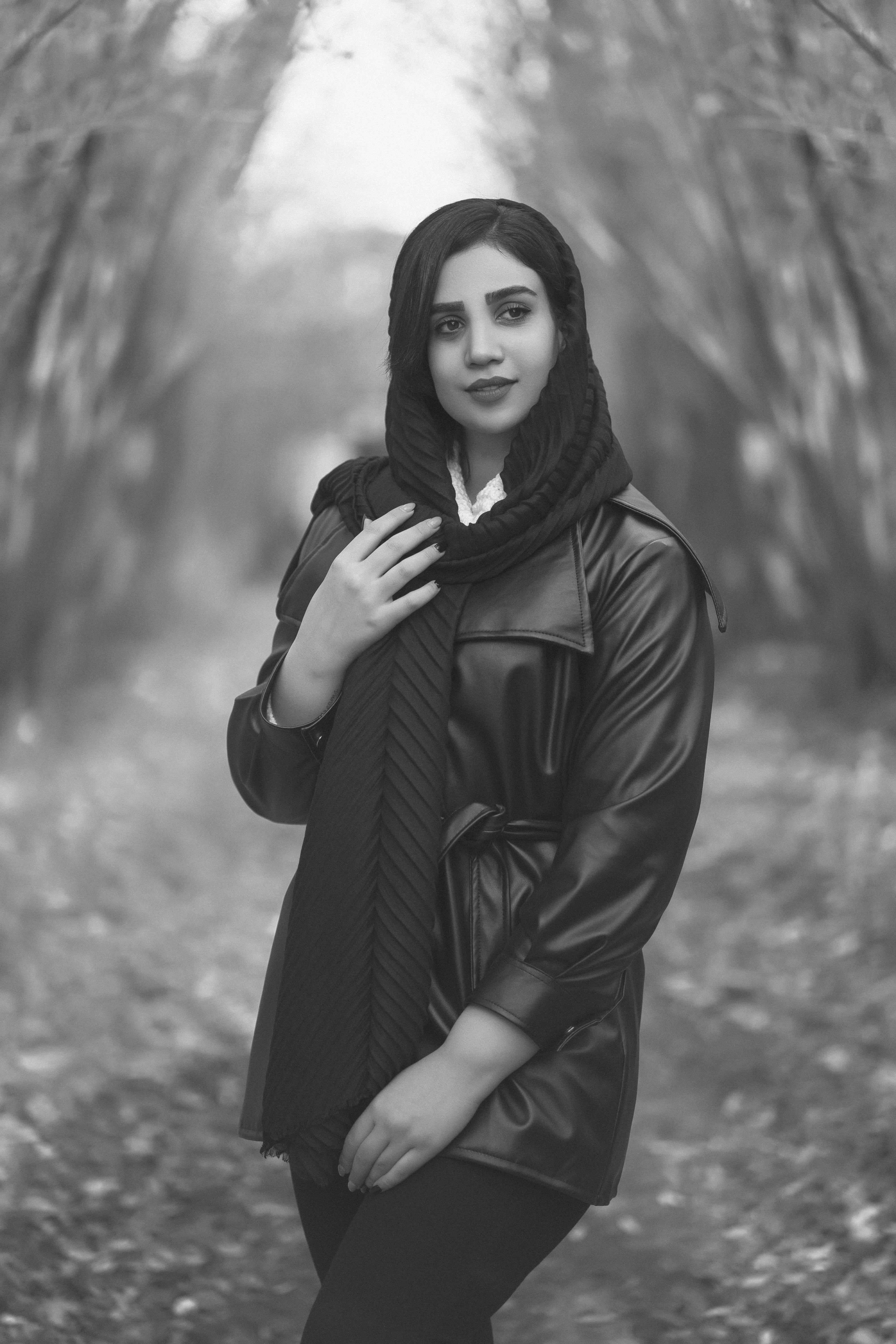 Black and white photo of a woman in a black leather jacket and headscarf posing in a forest in Hamedan.