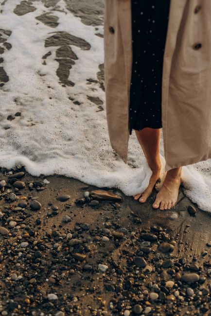 A woman in a coat stands barefoot on a pebble beach as waves gently reach the shore.